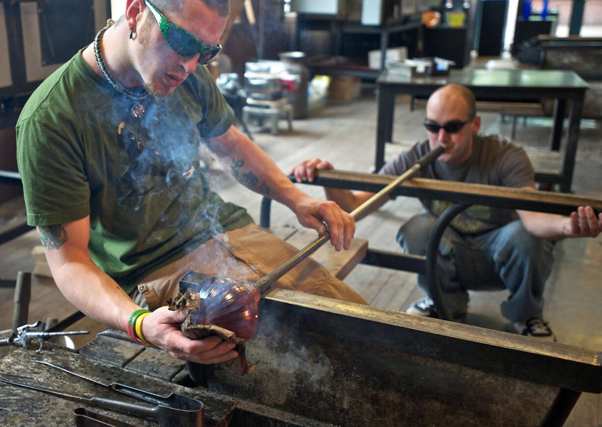 Andrew Lueck, manager of Firehouse Glass Studio, smooths and shapes the glass with newspaper while the shop tech Josh Handcock blows through the pipe.