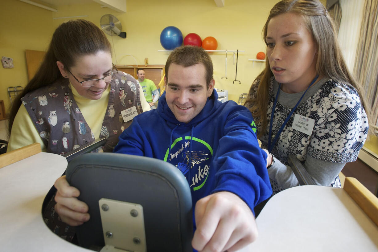 Luke Ashe is encouraged and lifted into a standing frame by Donelle Rodriguez, left, a physical therapy assistant, and Cassie Williams, a rehabilitation technician, during physical therapy Tuesday at Pacific Specialty and Rehabilitative Care in Vancouver. Luke stands, with help from the device, for 10 minutes to increase his endurance and strengthen his trunk muscles. Ashe was found in a coma Jan. 15.