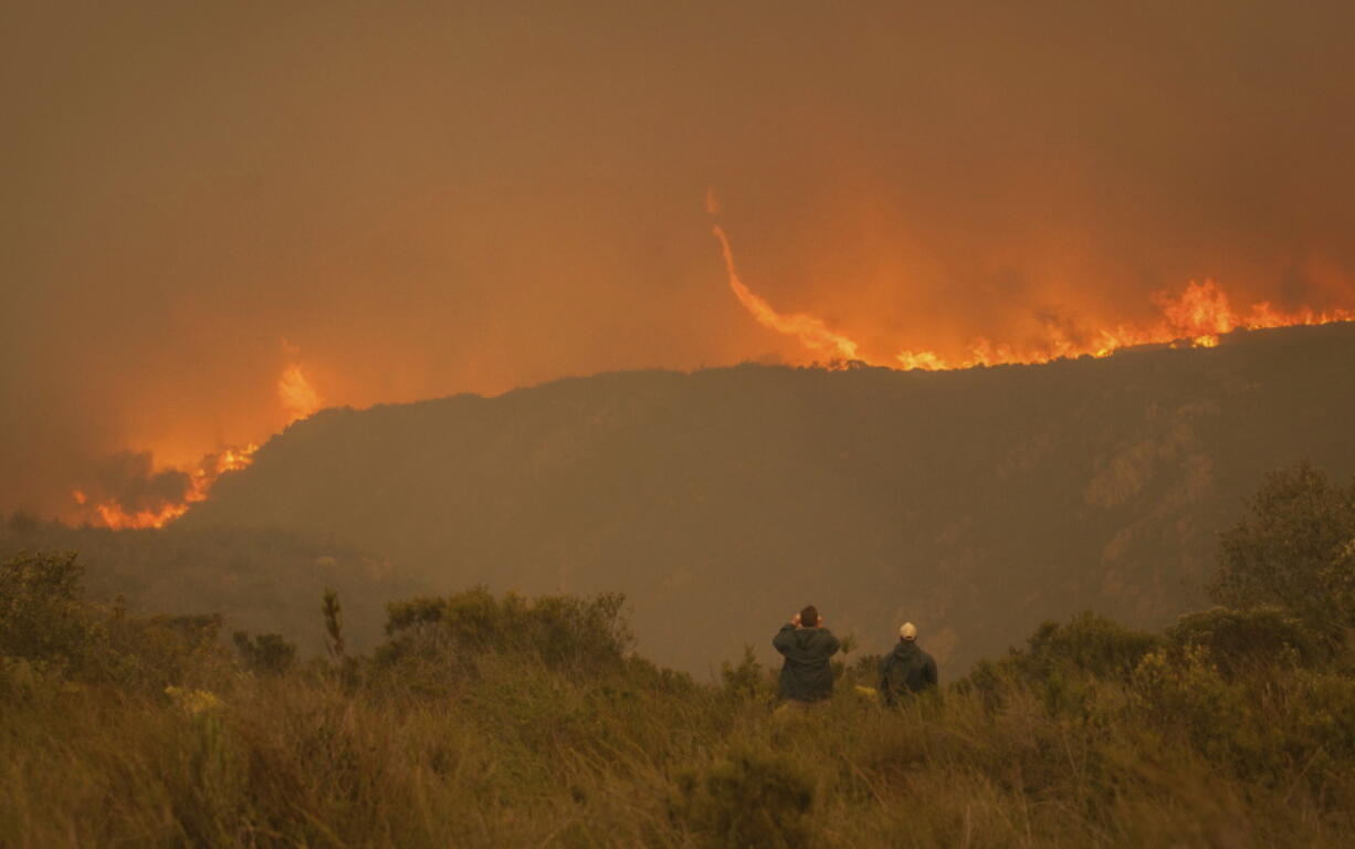 In this photo taken Wednesday, June 7, 2017, onlookers watch a blaze in the Kranshoek area of South Africa. The fire, fanned by high winds spread to nearby Plettenburg Bay and Knysna in the Western Cape Province killing several people, destroying homes and forcing the evacuation of up to 10,000 people.