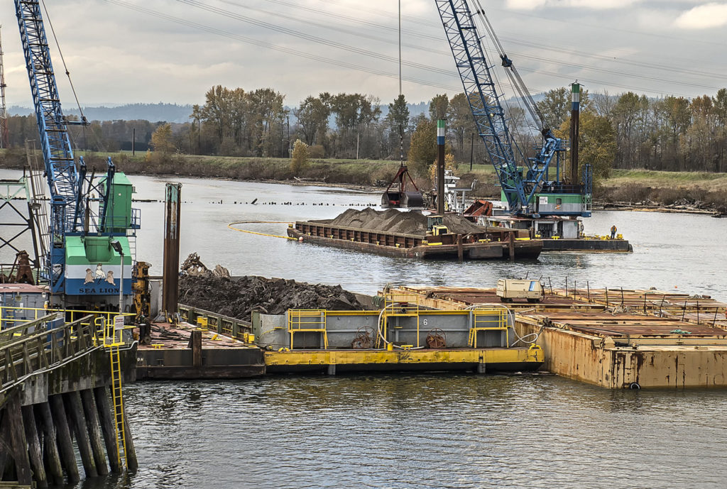 Legacy contamination from the former Reynolds aluminum plant is being cleaned up now just off the dike at Millennium Bulk Terminals Longview site on the Columbia River. Work started late last month dredging up a two-foot-thick layer of normal, silted river bottom and the two-foot-thick layer of contaminated soils which the silt covered. Wednesday, a barge-mounted crane was refilling the hole with certified clean sand. Work will continue today. The contaminated area is located just downriver from Millennium’s dock, and near where the company discharges all the treated surface water collected on the site back into the Columbia. The yellow-sided barge in the foreground contains some of the contaminated soil dredged earlier.