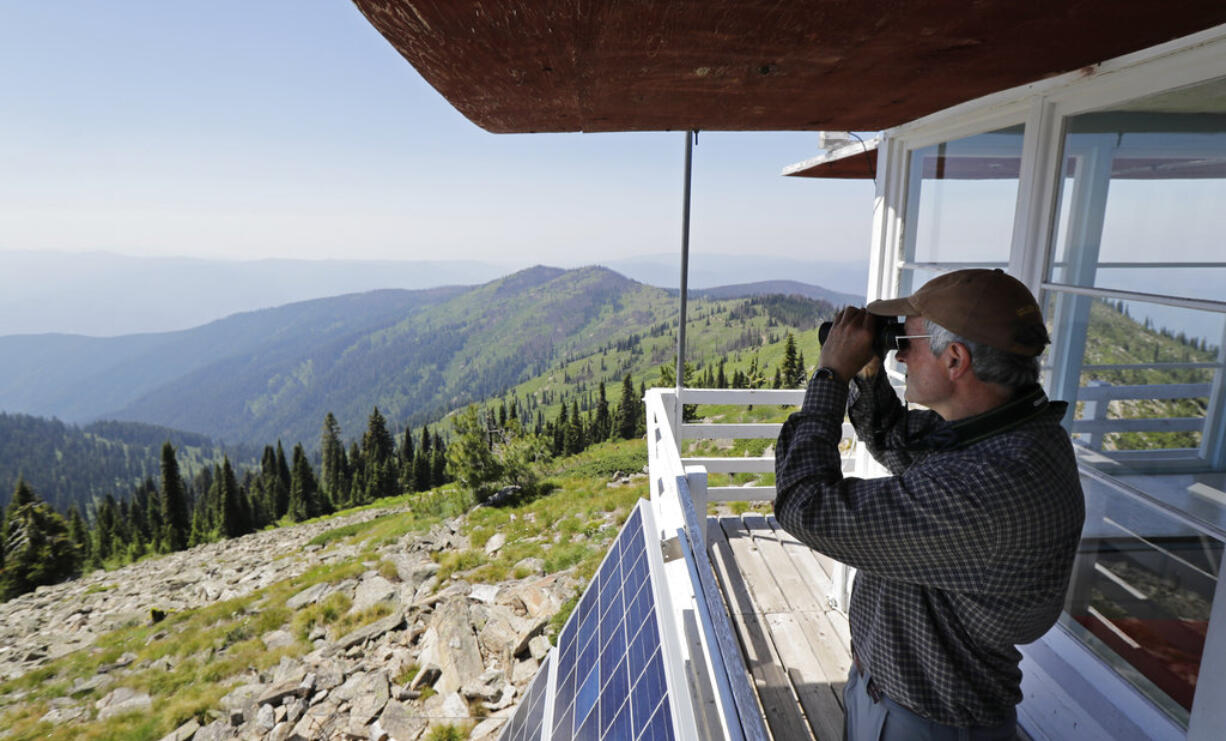 In this Wednesday, July 18, 2018, photo, Tom VandeWater stands at the railing of the Coolwater Fire Lookout and looks through his binoculars in the Nez Perce-Clearwater National Forests near Lowell, Idaho. VandeWater, from Canton, N.Y., has staffed the lookout each summer for many years for the U.S. Forest Service. Fire-lookout towers, perched atop remote, craggy peaks across the U.S. West, may seem like quaint reminders of an era before satellites, smartphones and jet-propelled air tankers, but the structures and the people who staff them play a crucial role in the nation's front-line efforts to stop wildfires. (AP Photo/Ted S.