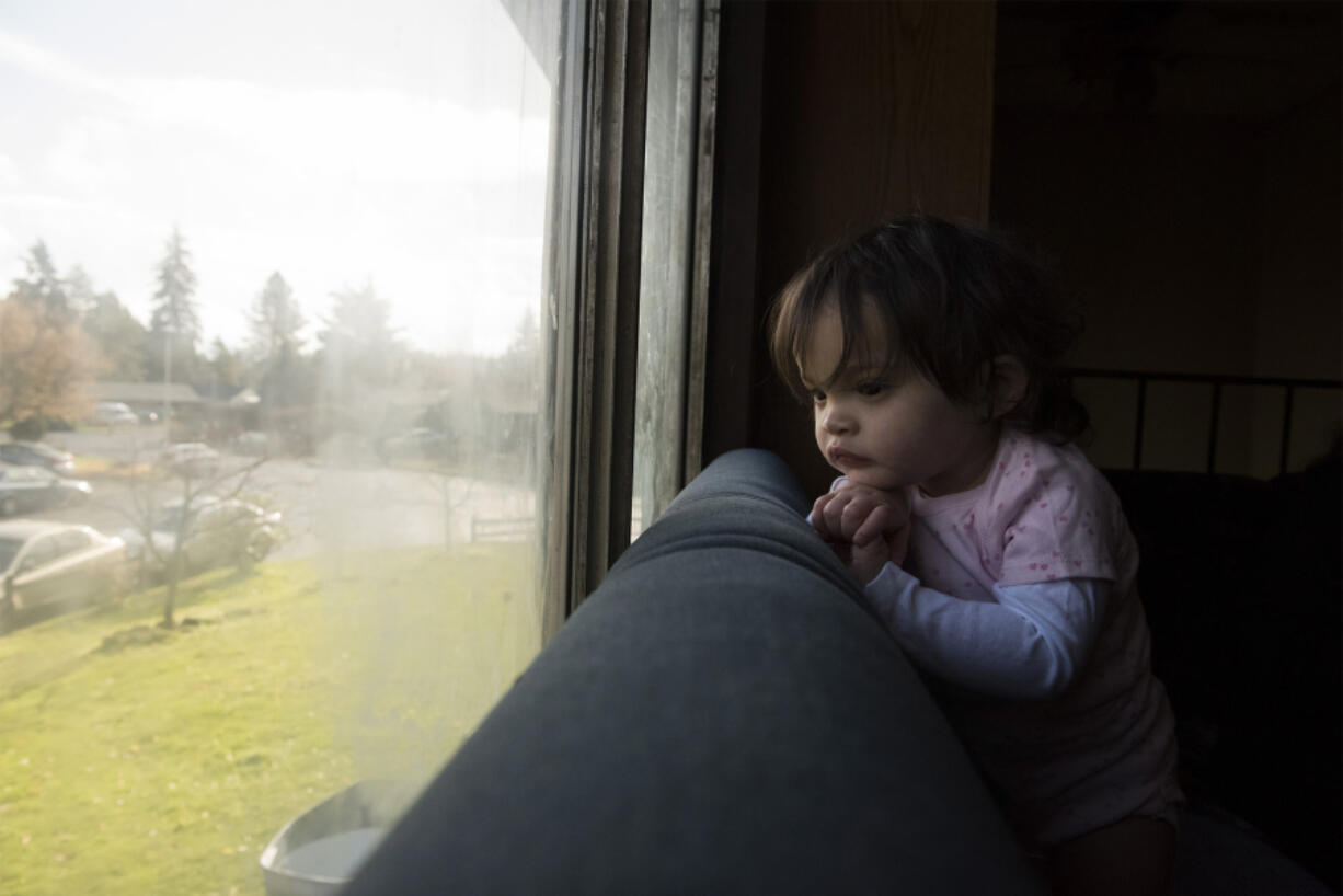 Kadence Sek, 1, watches her father, Isaac Sek, leave for work while she stays attached to her gastrointestinal tube feeding device at her home in Vancouver. Her mother, Cassie Sek, has had to cut hours at her job caring for mentally disabled adults to help care for Kadence. Isaac works six days a week as a carpenter, and regularly puts in 20 hours of overtime.