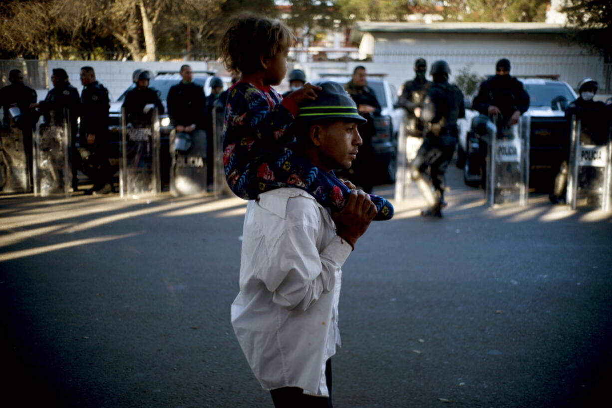 A migrant carries a child past Mexican police standing guard outside the Benito Juarez Sports Center, which is serving as a shelter for migrants in Tijuana, Mexico, Monday, Nov. 26, 2018. The mayor of Tijuana has declared a humanitarian crisis in his border city and says that he has asked the United Nations for aid to deal with thousands of Central American migrants who have arrived.