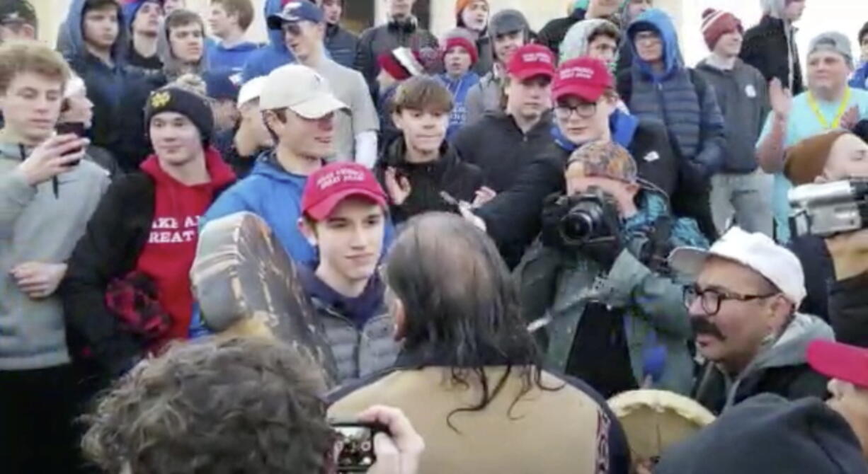 In this Friday, Jan. 18, 2019 image made from video provided by the Survival Media Agency, a teenager wearing a “Make America Great Again” hat, center left, stands in front of an elderly Native American singing and playing a drum in Washington. The Roman Catholic Diocese of Covington in Kentucky is looking into this and other videos that show youths, possibly from the diocese’s all-male Covington Catholic High School, mocking Native Americans at a rally in Washington.