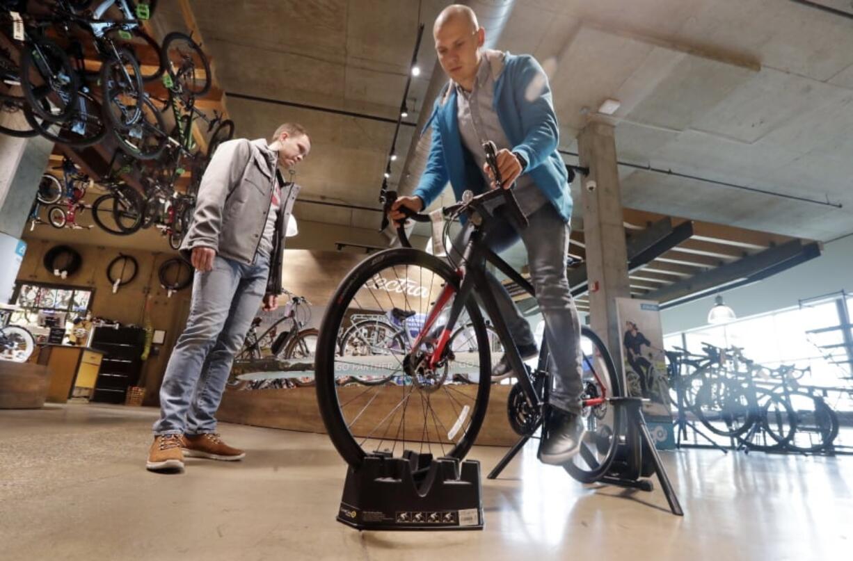 FILE- In this Nov. 13, 2018, photo Vitalii Myroshnychenko, right, test rides a bike atop a stand as Denis Rybalchenko looks on in REI Co-op’s flagship store in Seattle. The usual advice is “buy experiences, not things.” But that requires a deeper dive. A bicycle can provide an experience, and a new camera can preserve one. So buy experiences, especially with other people, but also think about buying material things that allow you to have experiences or enhance them.