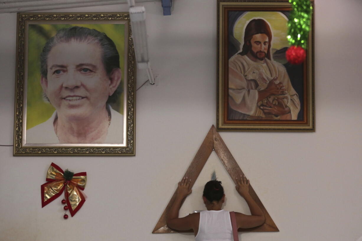 In this Jan. 4, 2019 photo, a woman places her hands in prayer on a wooden triangle, between framed images of spiritual healer Joao Teixeira de Faria and Jesus Christ, inside the Casa de Dom Inacio, in Abadiania, Brazil. Hundreds of women have accused de Faria of sexual abuse. The mounting accusations are turning the 77-year-old spiritual guru into Brazil’s first major figure to go down in the #metoo era.