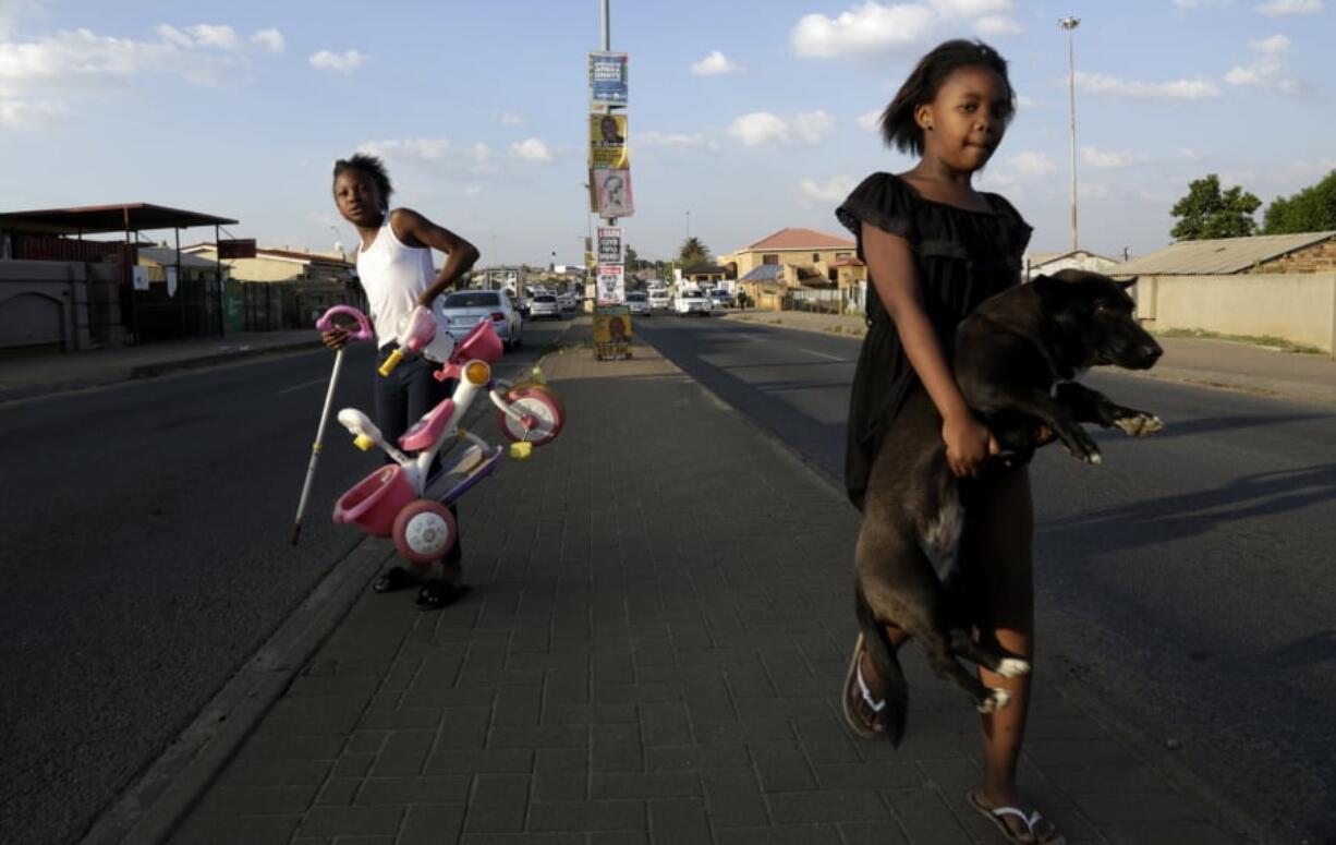 A young girl carries her dog as she crosses the main road, with headline from a newspaper and election posters on the streets of Soweto, South Africa, Friday, May 10, 2019. The ruling African National Congress is coasting to a comfortable lead in South Africa’s presidential and parliamentary elections with 80% of the vote counted, but the ongoing tally shows the party getting less support than in the previous poll five years ago.