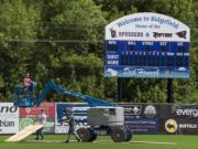 Mike Goldhammer of International Graphics & Nameplate, left, crosses the baseball field at Ridgefield Outdoor Recreation Complex while helping to install banners on the foul poles Monday . Crews put down boards as he crossed to protect the field.