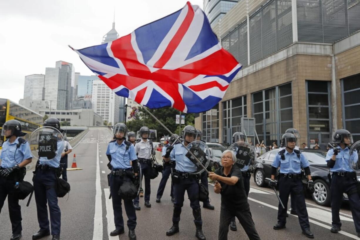 In this Wednesday, June 12, 2019, photo, a woman waves a British flag as policemen in anti-riot gear stand guard against the protesters on a closed-off road near the Legislative Council in Hong Kong. China promised that for 50 years after Britain gave up control of its last colony in 1997, this shimmering financial enclave would get to keep freedoms absent in the communist-ruled mainland that many here don’t want to live without.