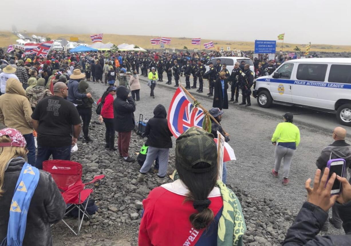 Police in riot gear are lined up on a road in Hawaii where an estimated 2,000 people are gathered to protest construction of a telescope on a mountain that some Native Hawaiians consider sacred, on Mauna Kea on the Big Island of Hawaii, Wednesday, July 17, 2019. Hawaii County Managing Director Wil Okabe says officials on Wednesday closed a highway leading to the protest site so a convoy of construction equipment can be brought up Mauna Kea. Protesters are off to the side of the road, singing.