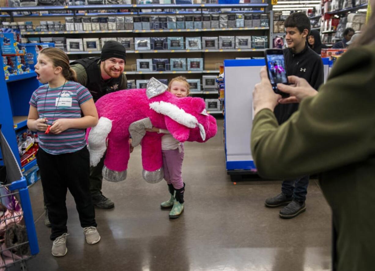 Faith Stanchfield, 6, center, smiles behind a plush, winged unicorn she picked out Saturday with Cowlitz County Sheriff&#039;s Deputy Craig Murray while sister Grace Stanchfield, left, ogles another toy during Woodland&#039;s annual &quot;Shop with a Cop&quot; event at Walmart in Woodland.