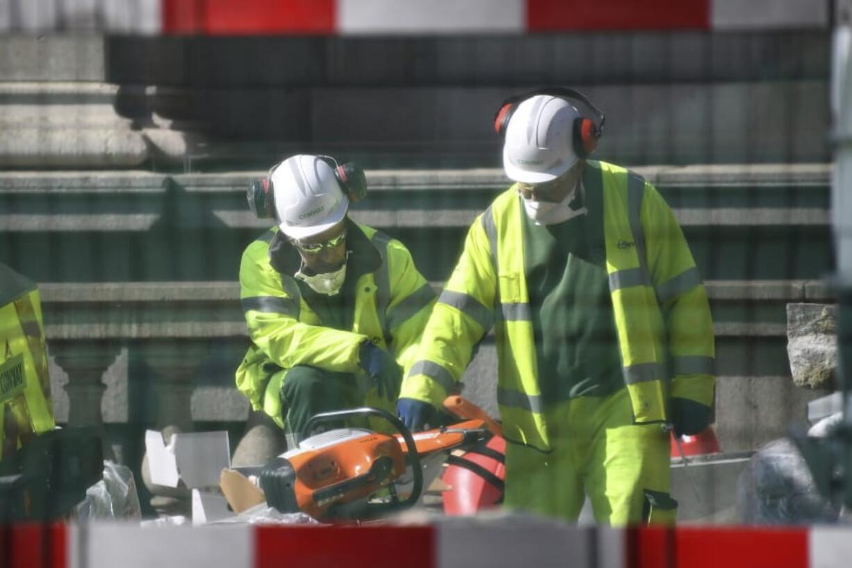 Construction workers wearing face masks seen through safety fencing work near London Bridge in London, Tuesday, March 24, 2020. Britain&#039;s Prime Minister Boris Johnson on Monday imposed its most draconian peacetime restrictions due to the spread of the coronavirus on businesses and gatherings, health workers begged for more gear, saying they felt like &quot;cannon fodder.&quot; For most people, the new coronavirus causes only mild or moderate symptoms. For some it can cause more severe illness.