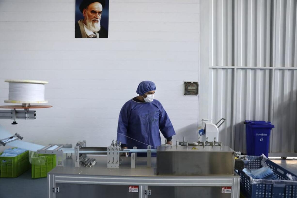 Under a portrait of the late Iranian revolutionary founder Ayatollah Khomeini, a worker wearing a protective suit controls a machine which makes face masks at a factory in Eshtehard, some 70 miles (114 kilometers) west of the capital Tehran, Iran, Tuesday, April 14, 2020. The factory is producing protective masks round-the-clock in a fight against the new coronavirus to meet the domestic demand as the country faces the worst outbreak in the Middle East.