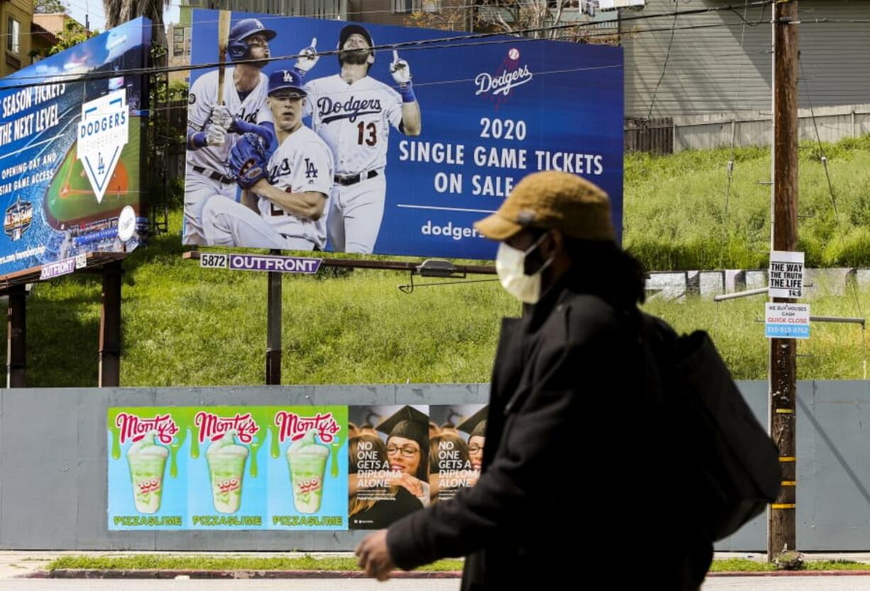 FILE - In this April 2, 2020, file photo, a pedestrian wears a hat and a face mask on Sunset Blvd., in the Echo Park neighborhood of Los Angeles. With no games being played, recent sports headlines have centered around hopes and dreams -- namely, the uncharted path leagues and teams must navigate to return to competition in the wake of the pandemic.