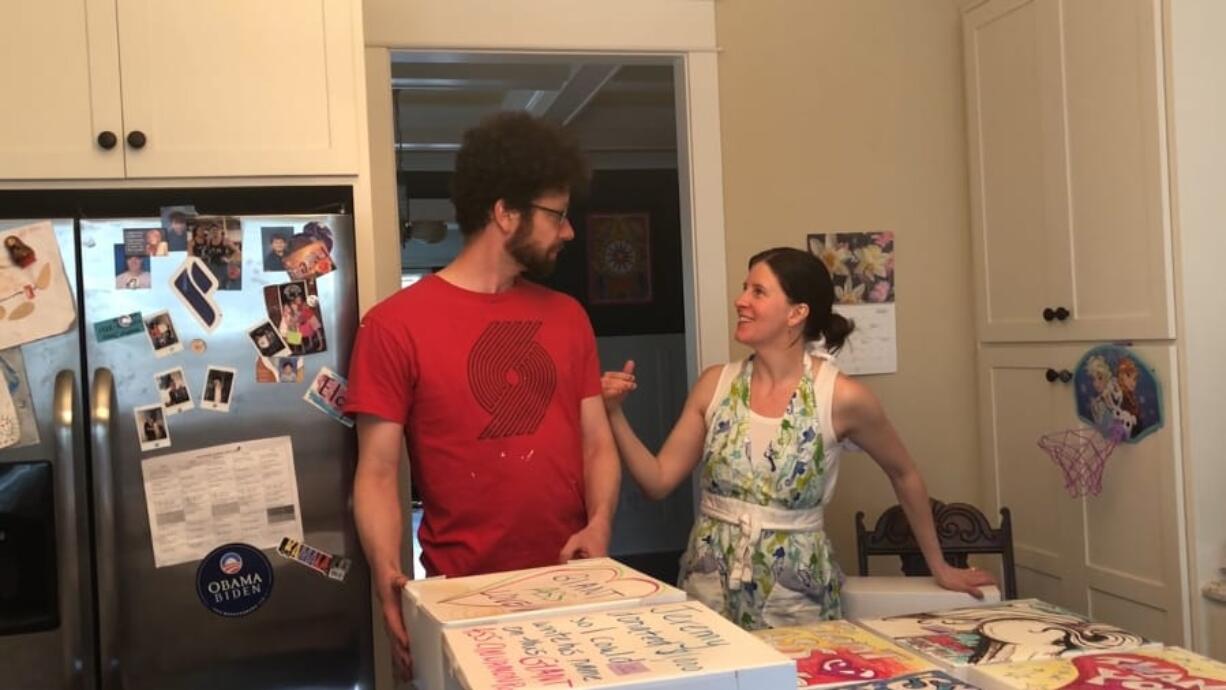 In this April 26, 2020, image from video provided by Whitney Rutz, Whitney Rutz, right, and husband, Paul, stand near several decorated boxes to hold large cinnamon rolls in their home in Portland, Ore. The Rutz family has helped raise thousands of dollars for Oregon Food Bank.