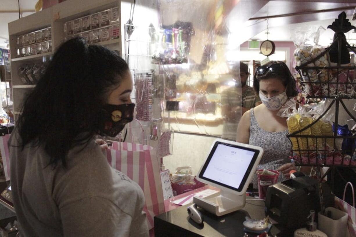 In this Thursday, May 28, 2020, photo, an employee at Bruce&#039;s Candy Kitchen ring up a customer&#039;s purchases from behind a protective plastic shield as both wear face masks due to the coronavirus in Cannon Beach, Ore. With summer looming, Cannon Beach and thousands of other small, tourist-dependent towns nationwide are struggling to balance fears of contagion with their economic survival in what could be a make-or-break summer.