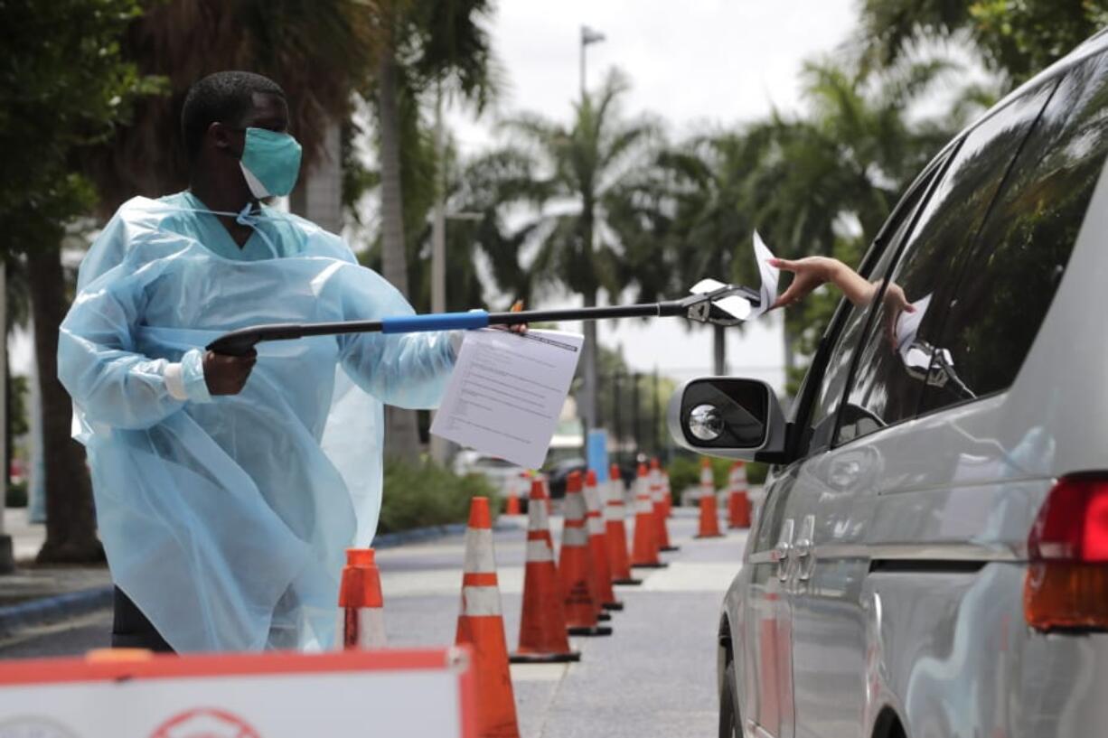 Healthcare worker Dante Hills, left, passes paperwork to a woman in a vehicle at a COVID-19 testing site outside of Marlins Park, Monday, July 27, 2020, in Miami. The Marlins home opener against the Baltimore Orioles on Monday night has been postponed as the Marlins deal with a coronavirus outbreak that stranded them in Philadelphia.