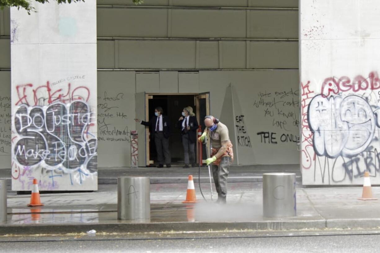 FILE - In this July 8, 2020, file photo, a worker washes graffiti off the sidewalk in front of the Mark O. Hatfield Federal Courthouse in downtown Portland, Ore., as two agents with the U.S. Marshals Service emerge from the boarded-up main entrance to examine the damage. Oregon&#039;s largest city is in crisis as violent protests have wracked downtown for weeks.