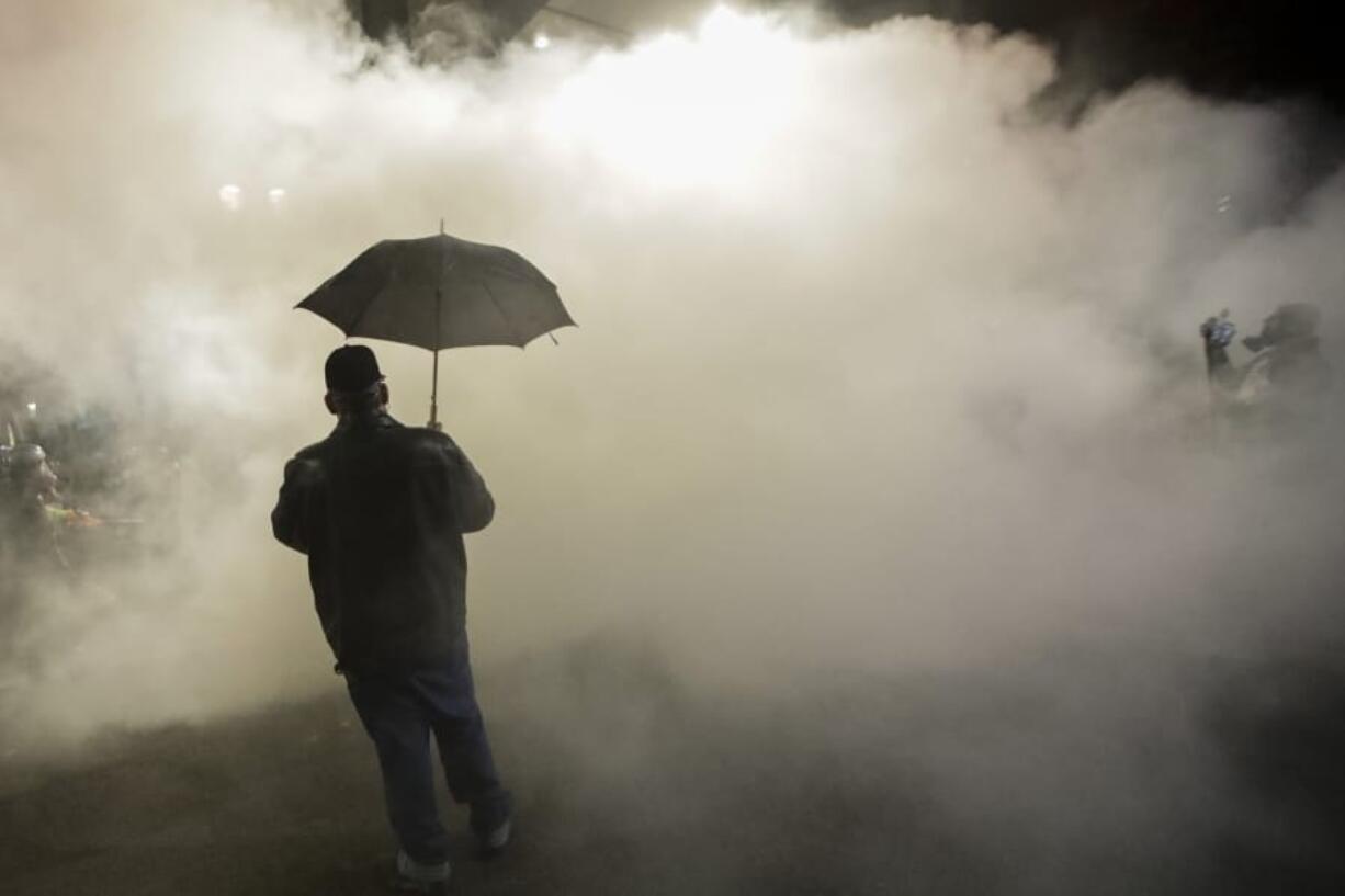 FILE - In this July 25, 2020, file photo, a protester carries an umbrella as federal police officers deploy tear gas during a protest at the Mark O. Hatfield U.S. Courthouse in Portland, Ore. Federal agents have left Portland, but city officials are still learning about and cleaning tear gas residue that lingers in the streets, dirt and possibly storm drains that empty into the Willamette River.