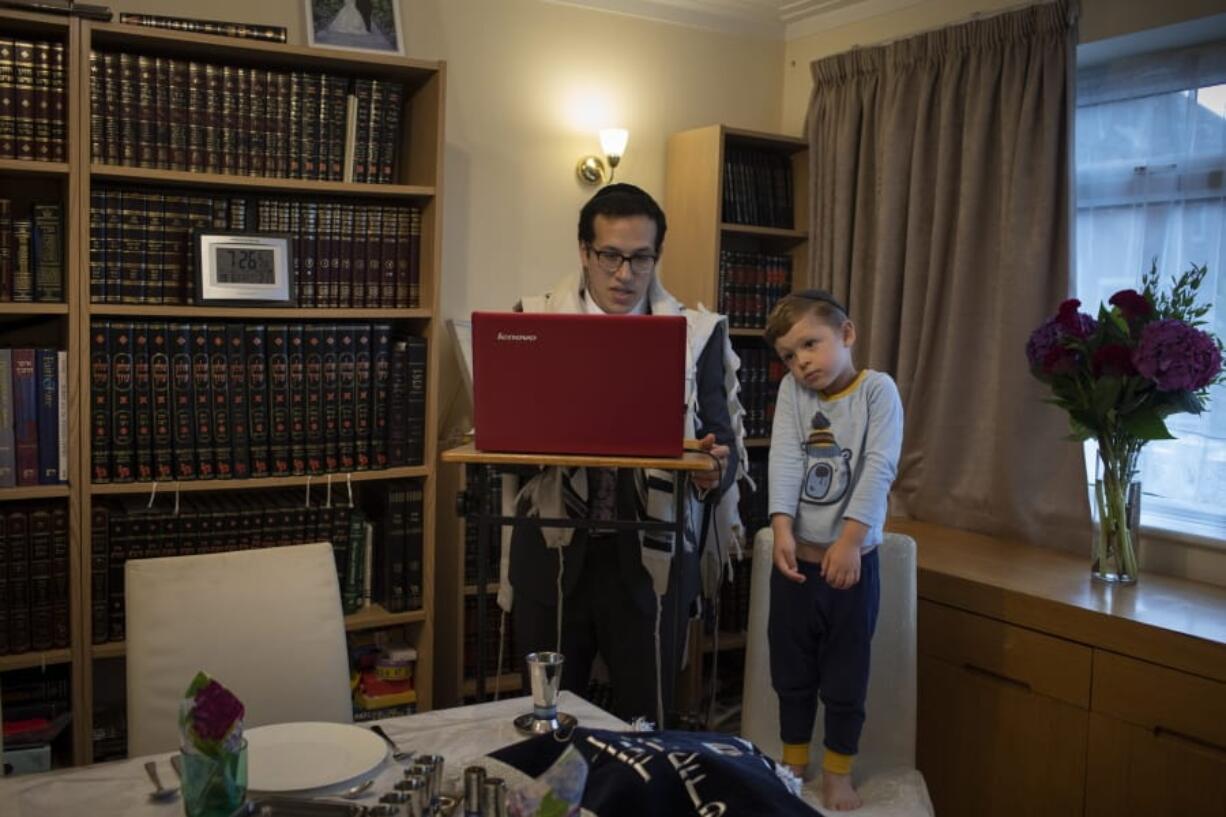 As his 3-year-old son Tzvi looks on, Rabbi Mordechai Chalk leads a service for his congregation via a teleconference app from his home in London on Friday, June 19, 2020, just before sunset. Taking services online-only has been particularly challenging for the Orthodox Jewish community, members of which are proscribed from using electronics on Shabbat, their day of rest.