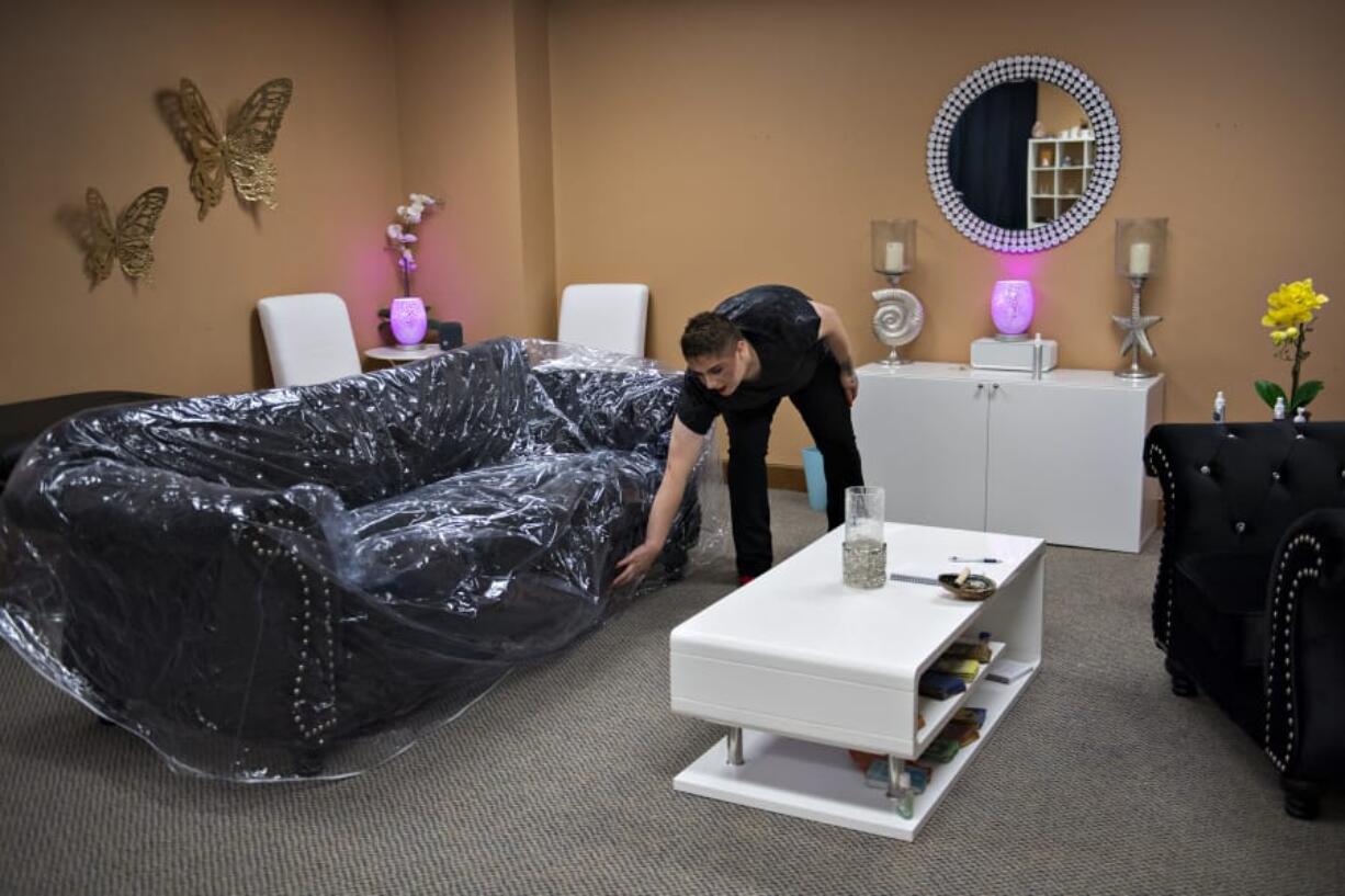 Daniel Monroe, 23, who works as a psychic medium in downtown Vancouver, wraps a couch in plastic that has been sanitized to protect against COVID-19 while preparing for an in-person client on a recent weekday. Monroe, who wears a mask during in-person sessions, also sees clients via Zoom.