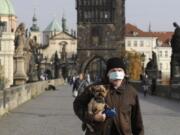 A man wearing a face mask carries his dog across the medieval Charles Bridge in Prague, Czech Republic, Wednesday, Oct. 21, 2020. In another desperate attempt to slow the rise of coronavirus infections in the Czech Republic, Health Minister Roman Prymula has announced a ban on free movement of people in the country and a closure of many stores, shopping malls and hotels. At the same time, state offices will limit their opening hours. Prime Minister Andrej Babis says those measures should prevent the collapse of the health system in early November.