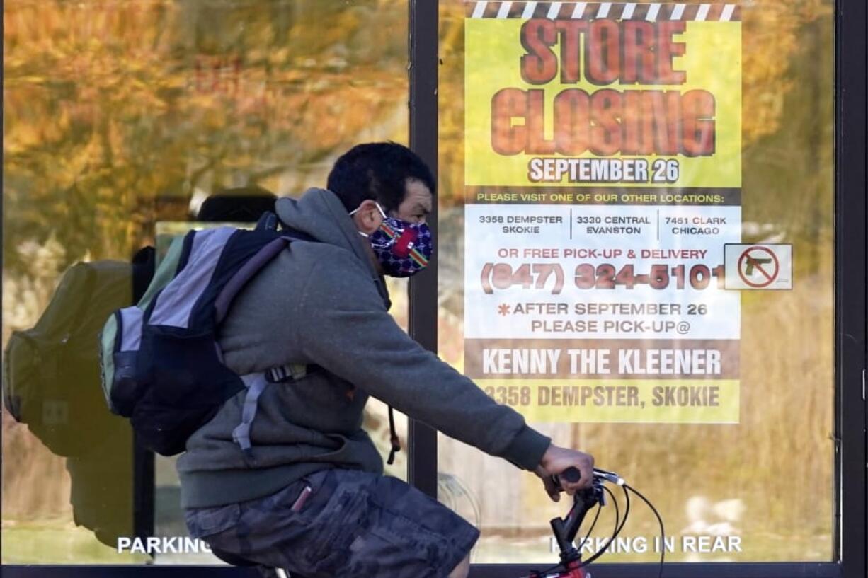 FILE - In this Friday, Nov. 6, 2020, file photo, a man wearing a face mask rides a bicycle past a closed store in Wilmette, Ill. States in the U.S. are renewing their push for more federal money to deal with the fallout from the coronavirus outbreak and to help them distribute a vaccine when one becomes widely available sometime in 2021. (AP Photo/Nam Y.