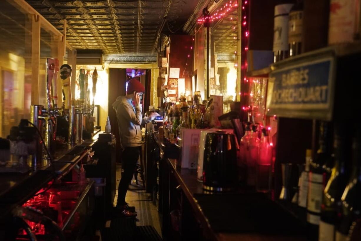 Bartender Alex Wright takes a phone order for liquor on his cell phone while tending the bar behind a protective plastic barrier separating him from patrons at Barbes, a popular music venue and bar converted to a bottle shop and mostly-outdoor service bar, Tuesday, Dec. 1, 2020, in New York.