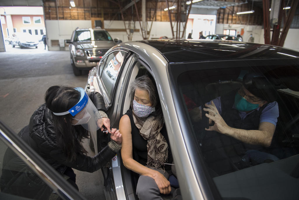 Kelly Nguyen of Safeway, left, gives a COVID-19 vaccination to Flor Bonura of Washougal as her husband, Thomas, snaps a photo with his smartphone at the Clark County Event Center at the Fairgrounds on Tuesday morning, January 26, 2021. Tuesday was the first day of mass vaccinations in Clark County, which are given by appointment only.