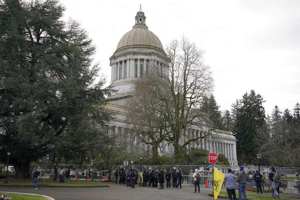 A group of anti-fascist protesters dressed in black make a brief stop outside a perimeter fence being watched by National Guard members and Washington State Patrol troopers, Sunday, Jan. 10, 2021, at the Capitol in Olympia, Wash. Washington Gov. Jay Inslee activated members of the National Guard this week to work with the Washington State Patrol to protect the Capitol campus. The group, part of a larger protest that was taking place in downtown Olympia, left after several minutes of exchanging words with troopers and guard members. (AP Photo/Ted S.