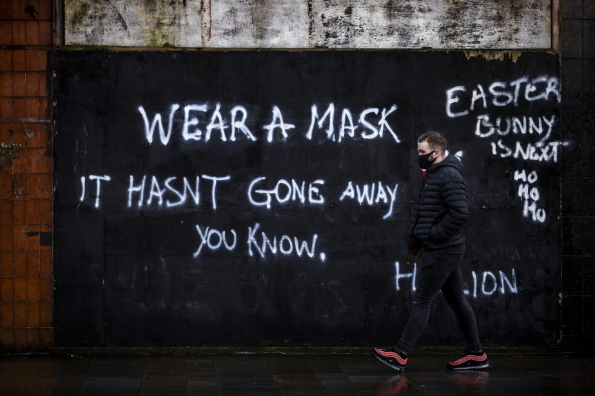 A man wearing a face covering walks past graffiti on the Lower Newtownards Road in Belfast with a message reading &#039;Wear a mask, it hasn&#039;t gone away you know&#039; Friday, Jan. 1, 2021.