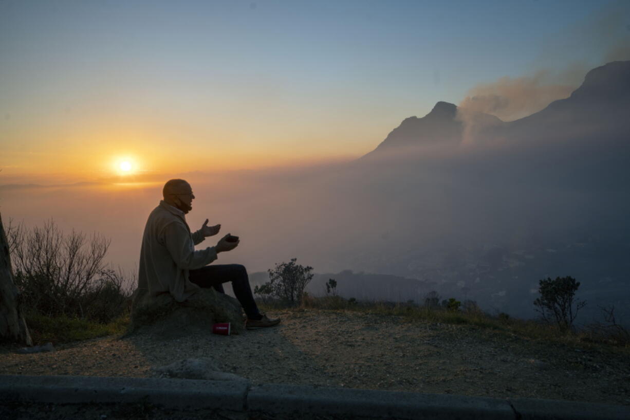 A Capetonian watches the sun rise from the top of Signal Hill as smoke engulfs the city of Cape Town, South Africa, Tuesday April 20, 2021. A massive fire spreading on the slopes of the city's famed Table Mountain, at right, is kept under control as firemen and helicopters take advantage of the low winds to contain the blaze.