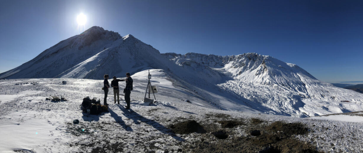 A field team from the Cascades Volcano Observatory discuss station maintenance plans on the north side of Mount St. Helens. Pictured in the center of the crater is a steaming lava dome from the 2004-08 eruption, and the fractured surface of Crater Glacier emerging from the gap on the north flank.