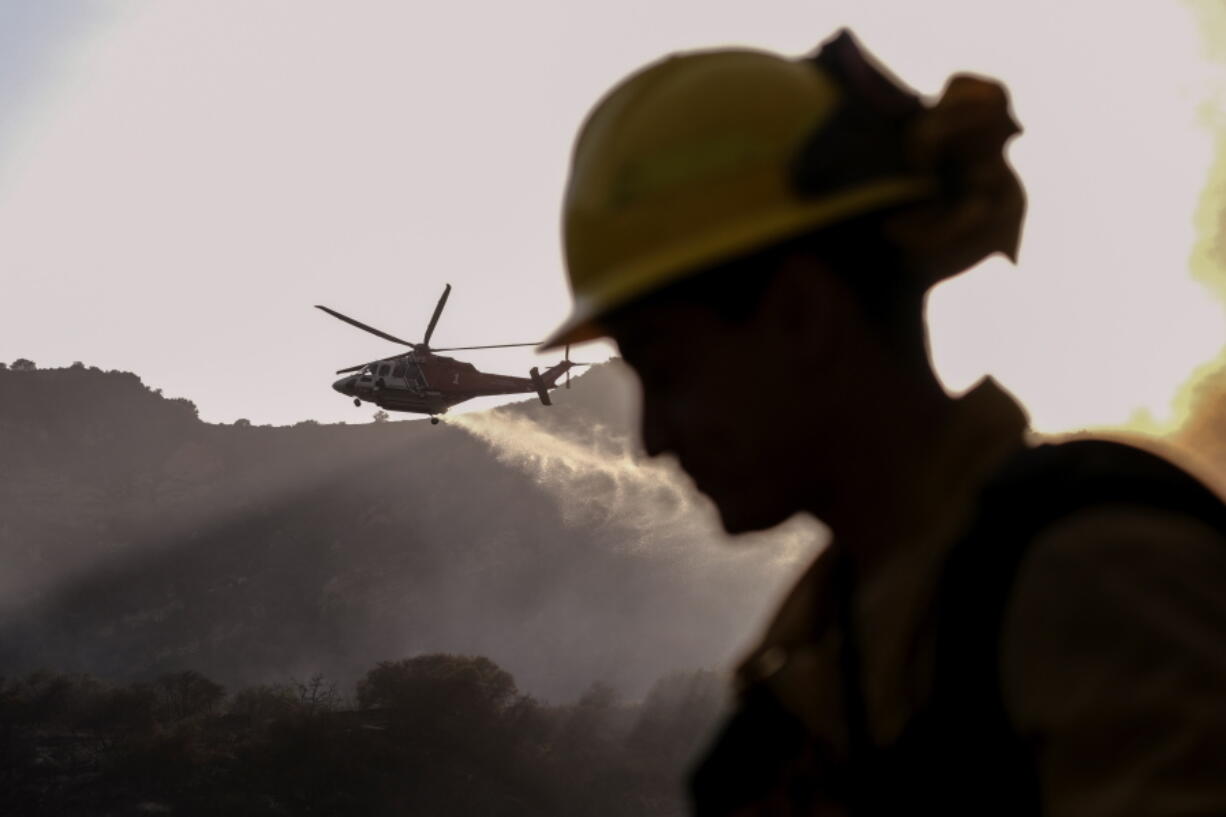 A firefighter keeps watch as a firefighting helicopter drops water on a brush fire scorching at least 100 acres in the Pacific Palisades area of Los Angeles on Saturday, May 15, 2021. (AP Photo/Ringo H.W.