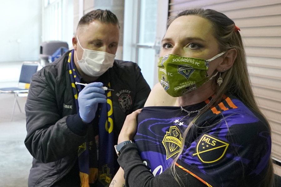 FILE - In this May 2, 2021, file photo, Stephanie Birman, right, a Seattle Sounders season ticket holder, wears a Sounders mask, jersey and earrings as she gets the Johnson & Johnson COVID-19 vaccine at a clinic in a concourse at Lumen Field prior to an MLS soccer match between the Sounders and the Los Angeles Galaxy. On Thursday, May 20, 2021, authorities in King County, Wash., which includes Seattle, said all people should continue wearing masks indoors until 70% of people in the county 16 and older are fully vaccinated. (AP Photo/Ted S.