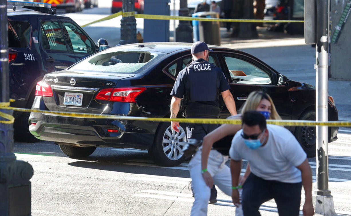 Pedestrians navigate crime scene tape, moving away from a car with windows shot as Seattle police are on the scene of a shooting in the Pioneer Square neighborhood, Sunday July 25, 2021, in Seattle. The unrelated shootings Sunday morning occurred in the Belltown, Pioneer Square, Chinatown International District and Capitol Hill neighborhoods.