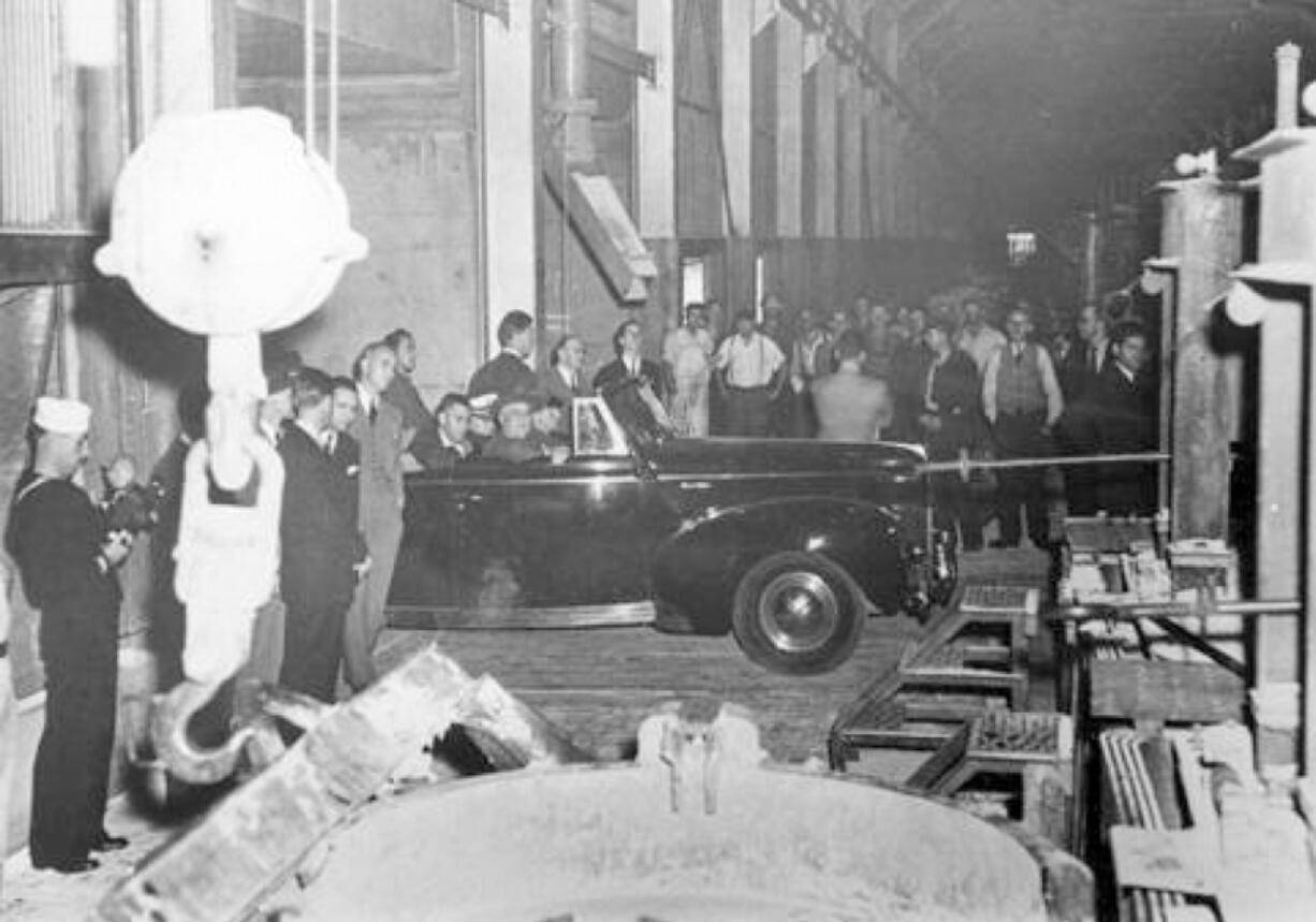 President Franklin D. Roosevelt quietly visited West Coast facilities contributing to the war effort in late 1942. He's shown in the convertible driving through the plant on the passenger's side with his arm resting on the door. He wanted to check on the status of manufacturing and aluminum critical to the war effort. In the left foreground is the crane used to lift huge vats. In the center is the lip of one of the used to hold molten aluminum. The men facing the crowd are likely Secret Service men protecting the president.