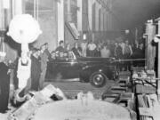 President Franklin D. Roosevelt quietly visited West Coast facilities contributing to the war effort in late 1942. He's shown in the convertible driving through the plant on the passenger's side with his arm resting on the door. He wanted to check on the status of manufacturing and aluminum critical to the war effort. In the left foreground is the crane used to lift huge vats. In the center is the lip of one of the used to hold molten aluminum. The men facing the crowd are likely Secret Service men protecting the president.