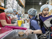 Volunteer Elyce Naray, right, carries her 1-year-old daughter, Elowen, on her back, as she helps package jambalaya meals at the Clark County Food Bank in Vancouver on Saturday. A $15,000 grant from The Church of Jesus Christ of Latter-day Saints to U.S. Hunger covered the cost of the food and packaging materials.