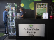 Budtender/cashier Robert Emmons, in blue mask, assists a customer as they pick up an online order inside Main Street Marijuana in Uptown Village on Tuesday morning. The store has put up new signs advising customers that all online orders must now be picked up at indoor counters following a change to state rules three weeks ago.
