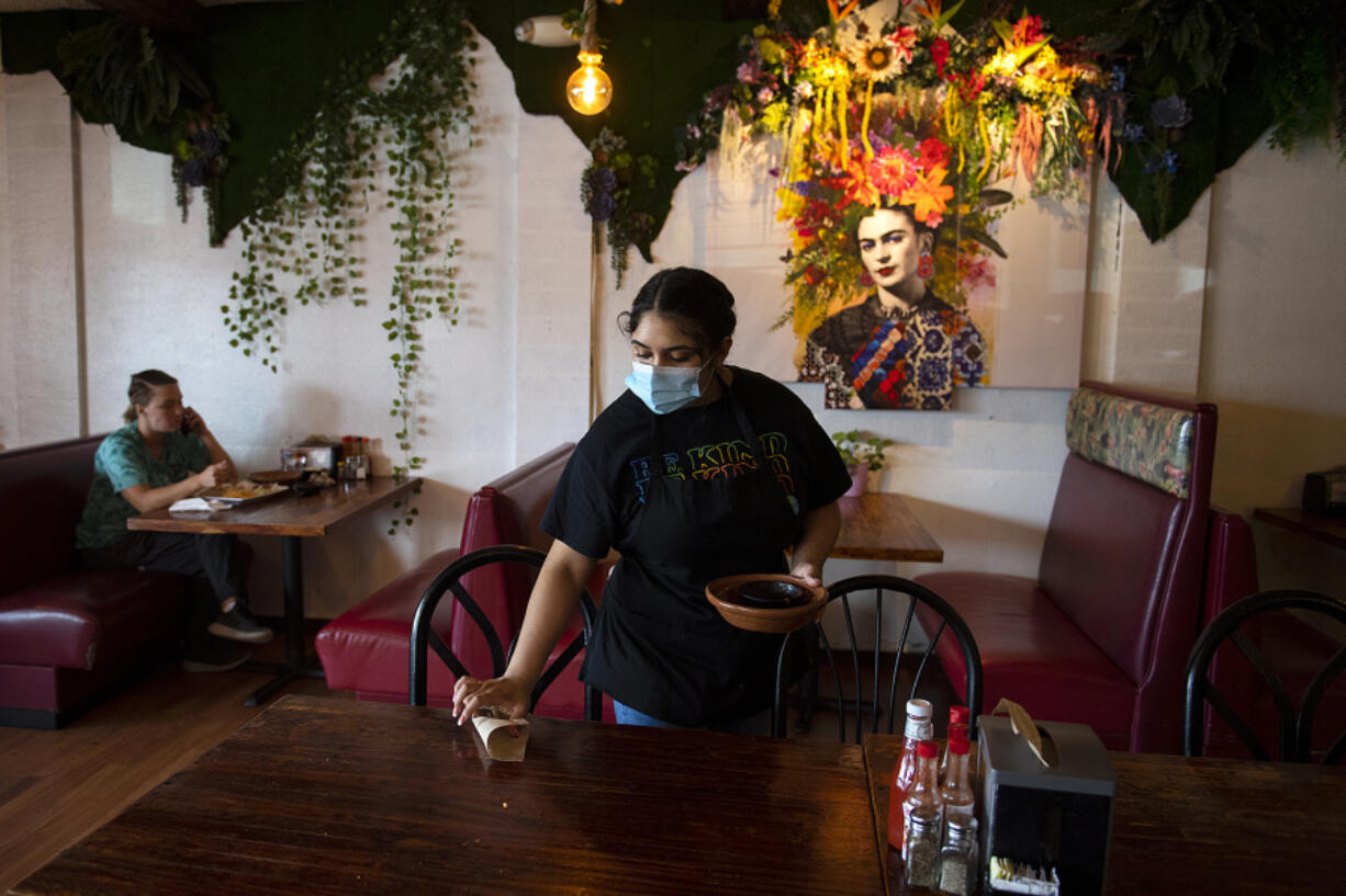 Vancouver resident Ashley Stevens enjoys lunch at 4 Caminos as Samantha Gomez cleans up after lunch customers on a recent Wednesday. Hispanic restaurants on Fourth Plain have had challenges with rising supply costs and labor shortages. At top, a sign at 4 Caminos lets customers know masks are required inside.