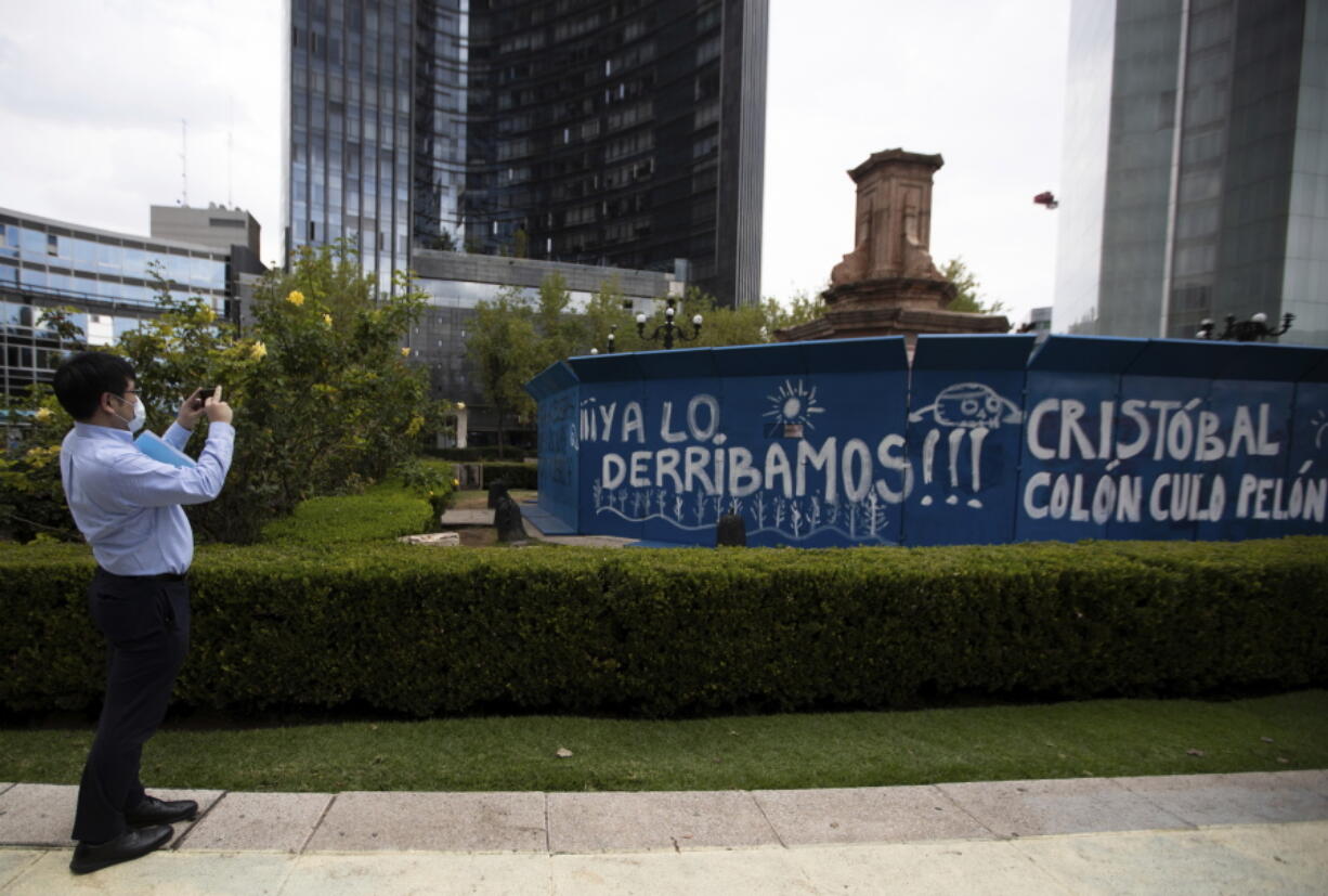 FILE - In this Oct. 12, 2020 file photo, a pedestrian takes a photo of graffiti on a temporary metal barrier set up to protect the perimeter of the Christopher Columbus's statue which was removed by authorities on Paseo de la Reforma in Mexico Cito. Mexico City Mayor Claudia Sheinbaum announced on Sunday, Sept. 5, 2021 that the statue will be replaced by a statue honoring Indigenous women.