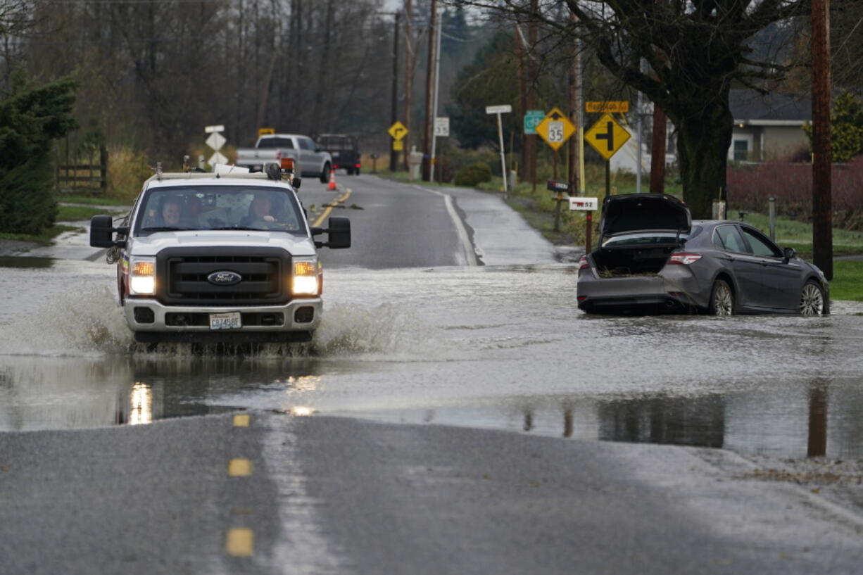 A truck drives through water over a road near Everson, Wash., Monday, Nov. 29, 2021 past a car that was stranded by flooding in the area earlier in the month. Localized flooding was expected Monday in Washington state from another in a series of rainstorms, but conditions do not appear to be as severe as when extreme weather hit the region earlier in November.