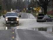 A truck drives through water over a road near Everson, Wash., Monday, Nov. 29, 2021 past a car that was stranded by flooding in the area earlier in the month. Localized flooding was expected Monday in Washington state from another in a series of rainstorms, but conditions do not appear to be as severe as when extreme weather hit the region earlier in November.