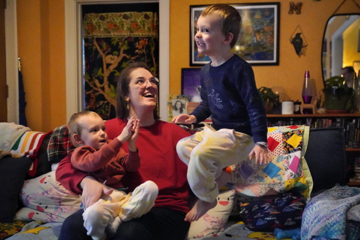 Heather Cimellaro holds her three-year-old son Charlie while his twin brother, Milo, jumps on a couch at their home, Wednesday, Jan. 5, 2022, in Auburn, Maine. Heather Cimellaro is one many parents concerned about the omicron surge and the dilemma it's posing for families of children too young to be vaccinated. (AP Photo/Robert F.