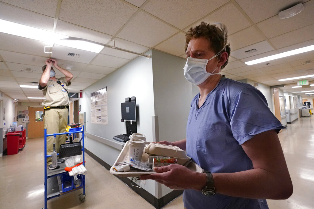 Carpenter Shayne Pitt, left, works to place brackets for curtains to add space for patient beds in a hallway as registered nurse Scott McGieson carries a meal to a patient's room in the acute care unit of Harborview Medical Center, Friday, Jan. 14, 2022, in Seattle. Washington Gov. Jay Inslee is deploying 100 members of the state National Guard to hospitals across the state amid staff shortages due to an omicron-fueled spike in COVID-19 hospitalizations. Inslee announced Thursday that teams will be deployed to assist four overcrowded emergency departments at hospitals in Everett, Yakima, Wenatchee and Spokane, and that testing teams will be based at hospitals in Olympia, Richland, Seattle and Tacoma.