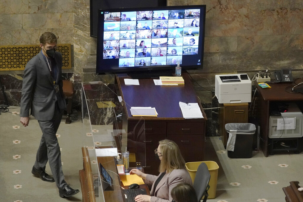 With most Senators attending remotely and shown on a video display, legislative workers are shown on the Senate floor, Wednesday, Jan. 26, 2022, at the Capitol in Olympia, Wash., during debate on a measure that would delay implementation of a long-term care program and the payroll tax that pays for it. The Senate passed the measure, which was passed by the House last week, and Gov. Jay Inslee is expected to sign the measure on Friday. (AP Photo/Ted S.