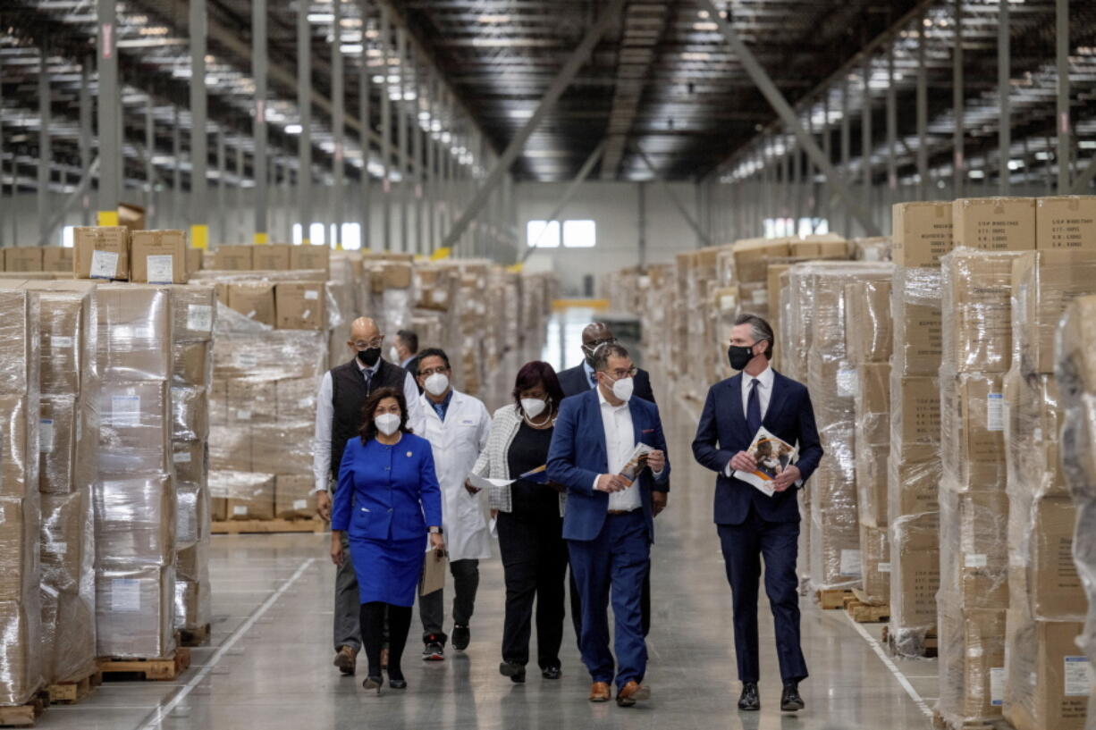 Gov. Gavin Newsom, right, walks through rows of boxed Personal protective equipment, PPE, with dignitaries and elected officials, as he prepares to announce the next phase of California's COVID-19 response called "SMARTER," during a press conference at the UPS Healthcare warehouse in Fontana, Calif., on Thursday, Feb. 17, 2022. The plan is to move from the pandemic stage into an endemic stage in which people will learn to live COVID.