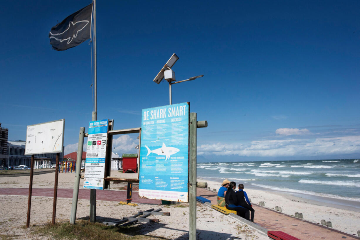 People sit next to shark-education signs, and the shark-warning flag at Muizenberg beach, which is part of False Bay in Cape Town, South Africa.