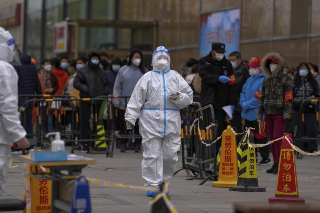 A health worker wearing a protective suit walks by masked residents who wait in line to get their throat swab at a coronavirus testing site following a COVID-19 case was detected in a residential buildings, Wednesday, April 6, 2022, in Beijing.